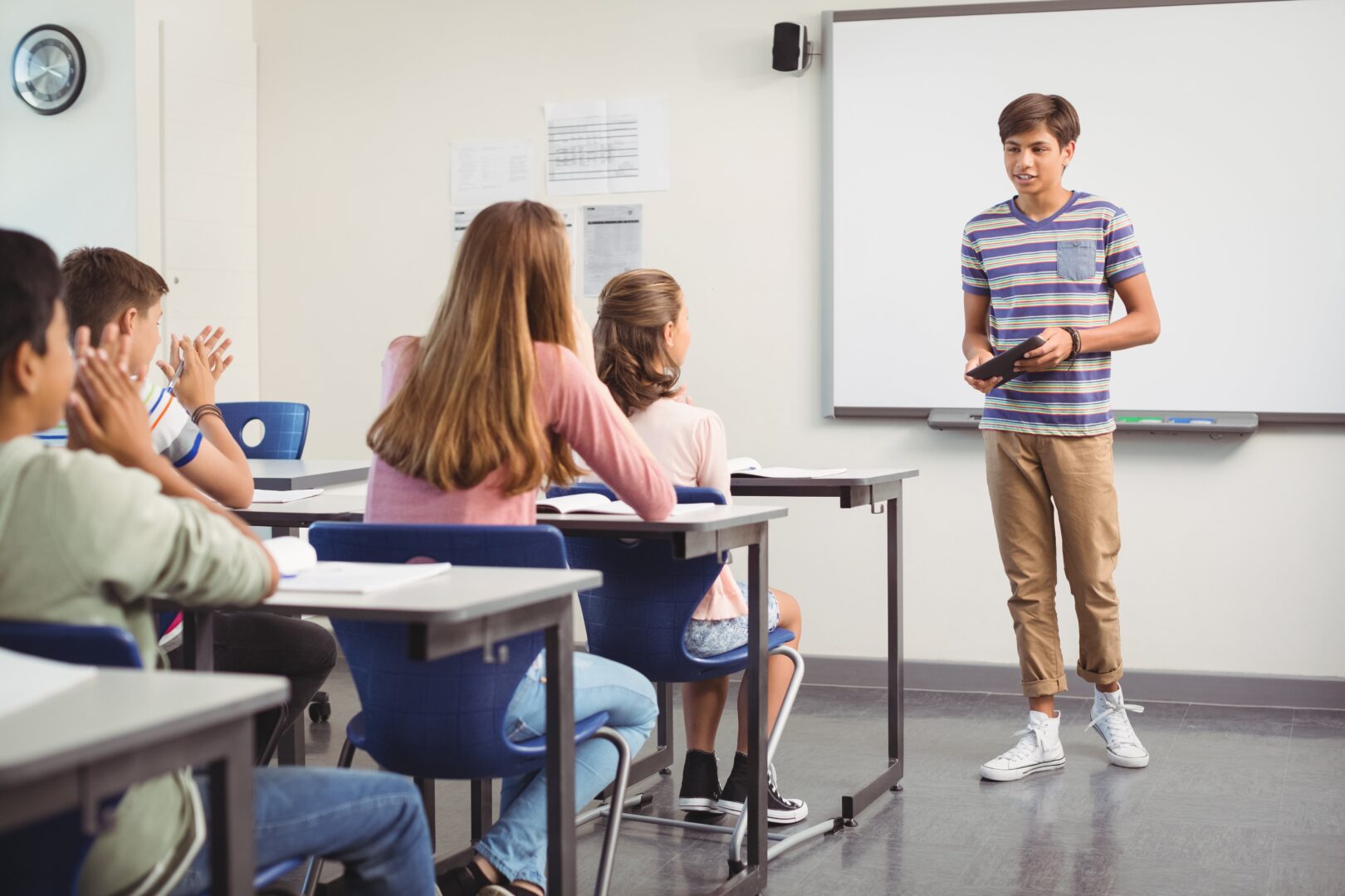 A teen presenting to a small group of classmates at a whiteboard, peers applauding from their desks